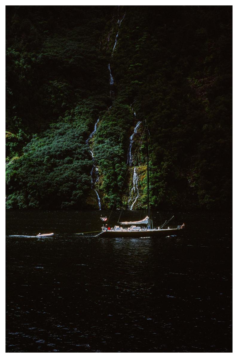 A sailboat and a small dinghy are on dark water with a lush green mountain and waterfalls in the background.