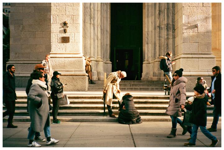 People walk on a sunny street in front of a large stone building with a grand entrance.