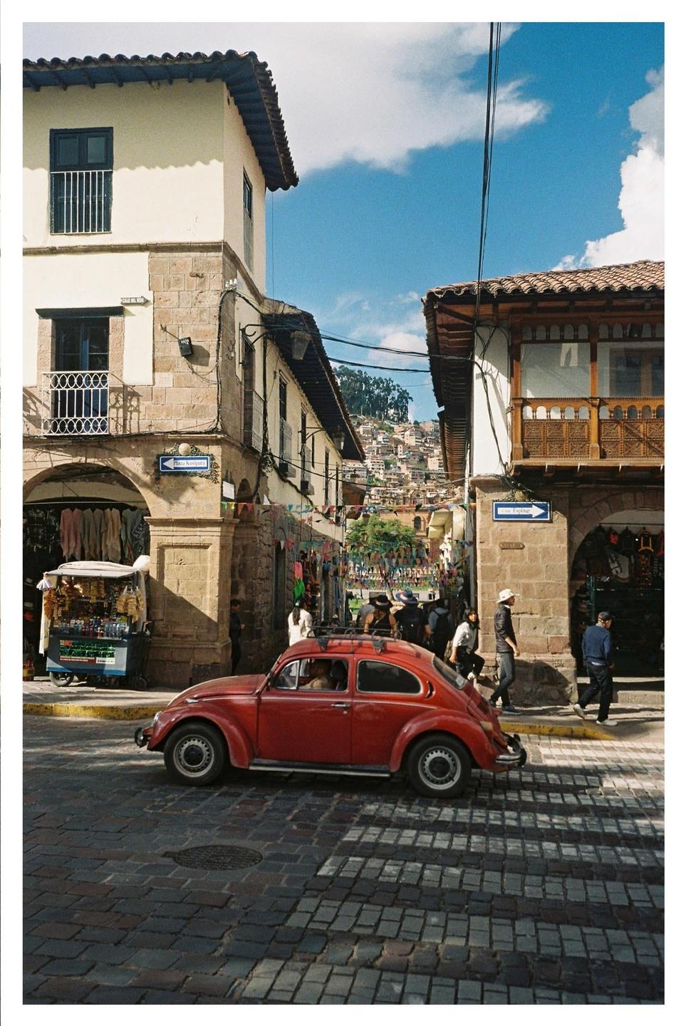 A red car is parked on a cobblestone street in a town with buildings and people.