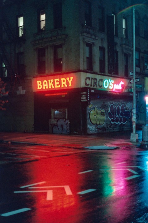 A neon-lit bakery and pastry shop with graffiti on its facade reflects onto a wet street at night.
