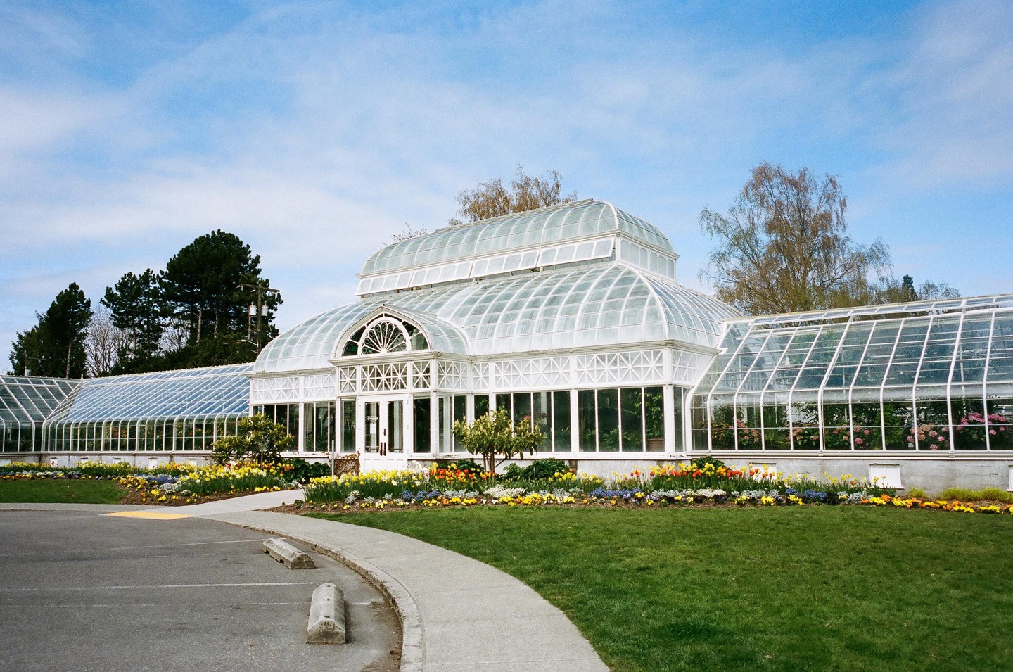 A large white conservatory with a glass roof is surrounded by green grass and colorful flowers under a blue sky.