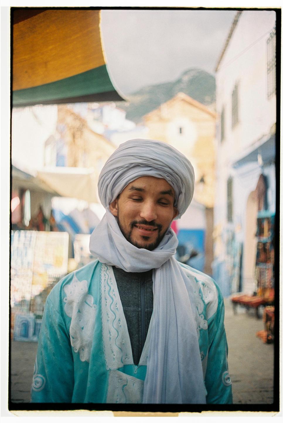 A man wearing a light blue turban and robe smiles with his eyes closed on a street.
