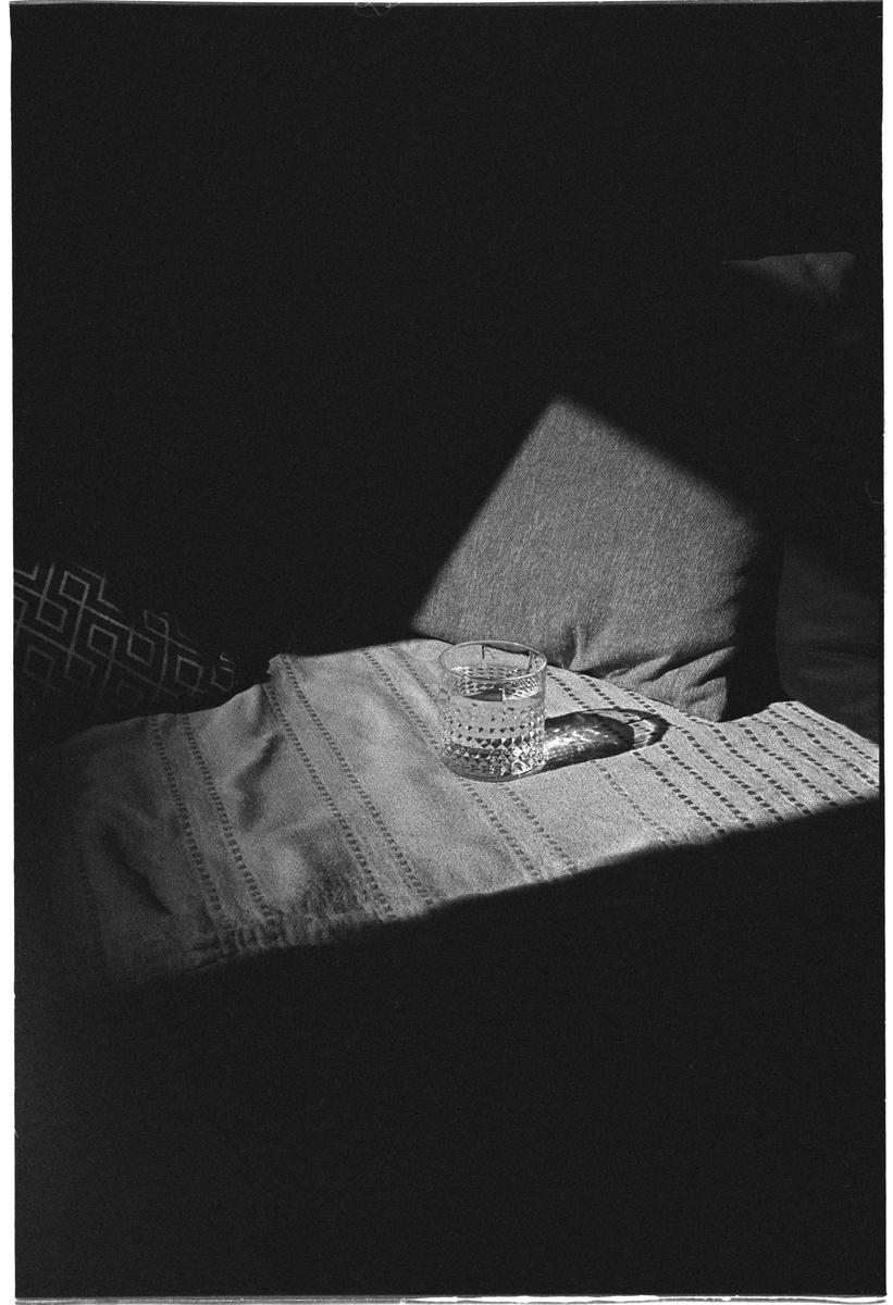 A glass of water sits on a patterned blanket with a pillow in the background, all partially illuminated by a strong diagonal beam of light.