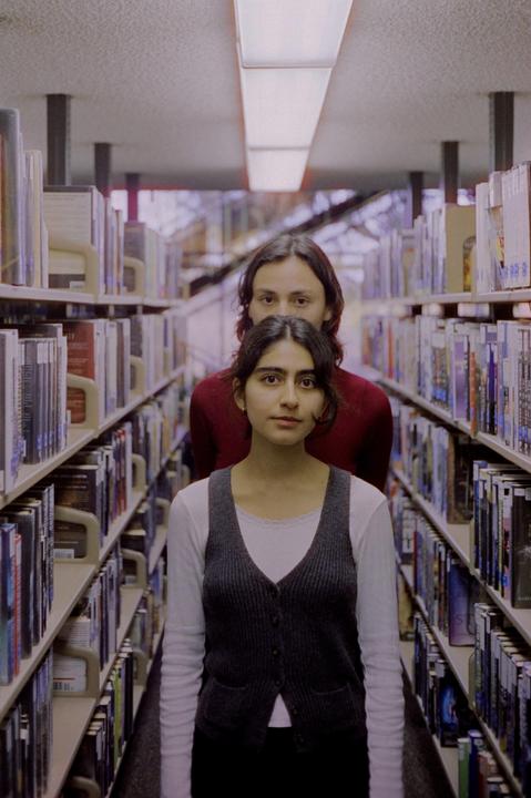 A woman stands in front of a man in a library aisle, both looking directly at the viewer.