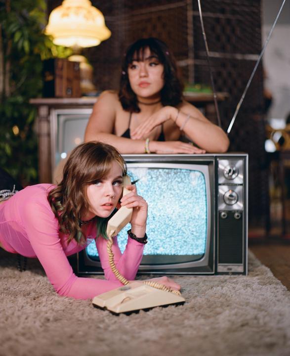 A woman lies on a carpet holding a telephone receiver while another woman leans on a television set behind her.