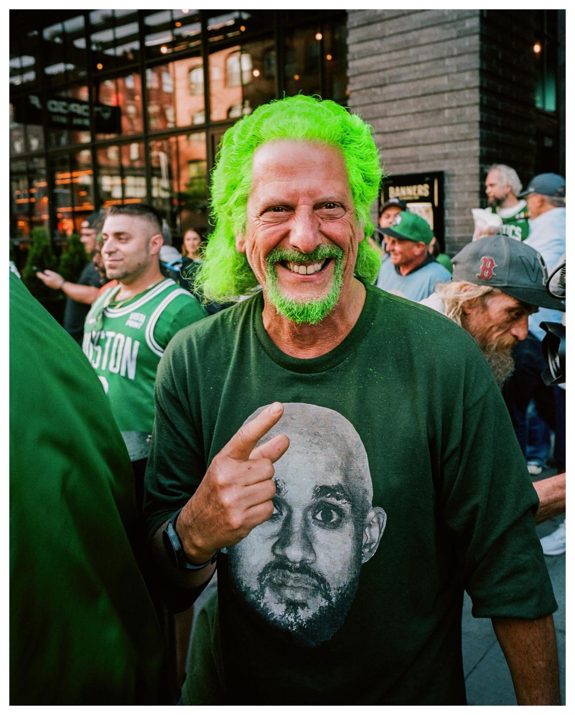 A man with bright green hair and beard smiles at the camera while wearing a green t-shirt with a face printed on it.