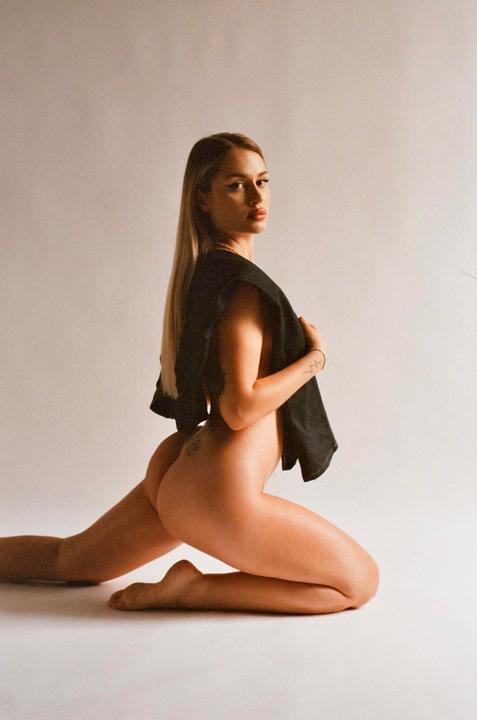 A woman kneels on a white surface, looking over her shoulder at the viewer.