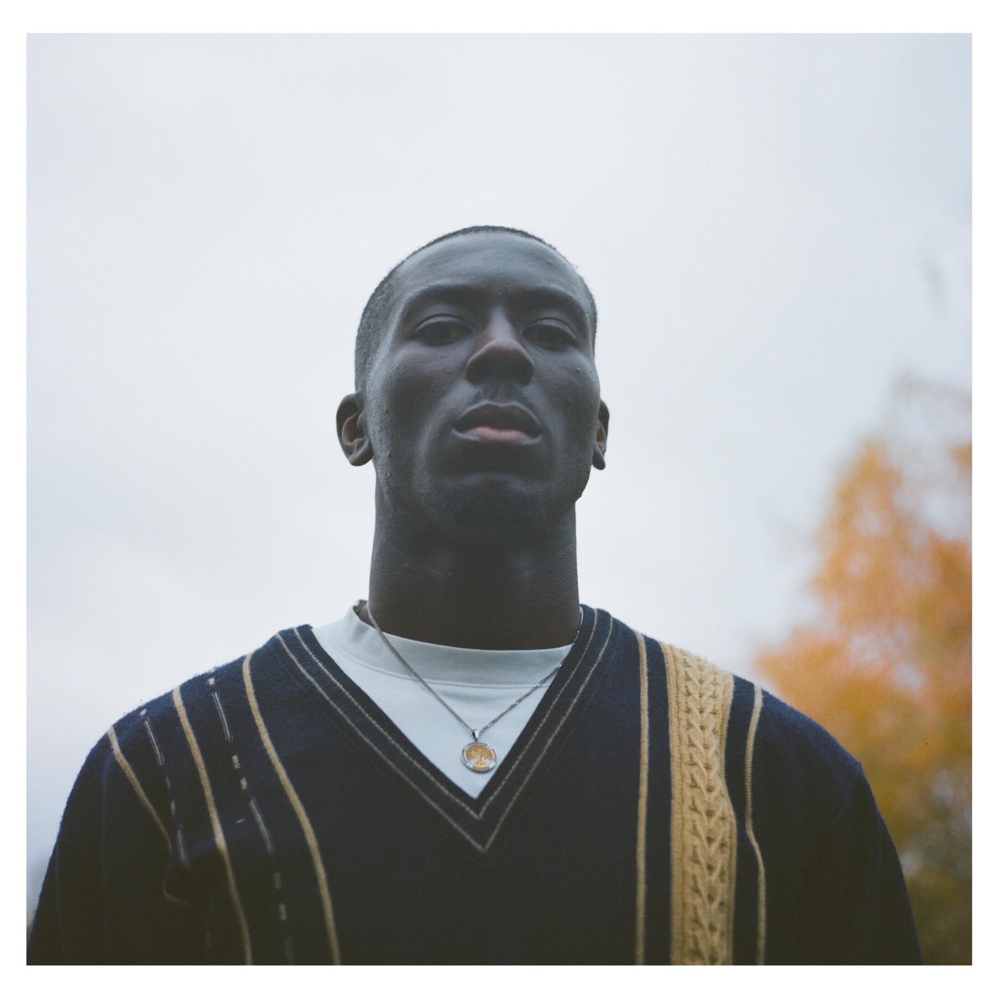 A man wearing a sweater and a necklace looks directly at the viewer.