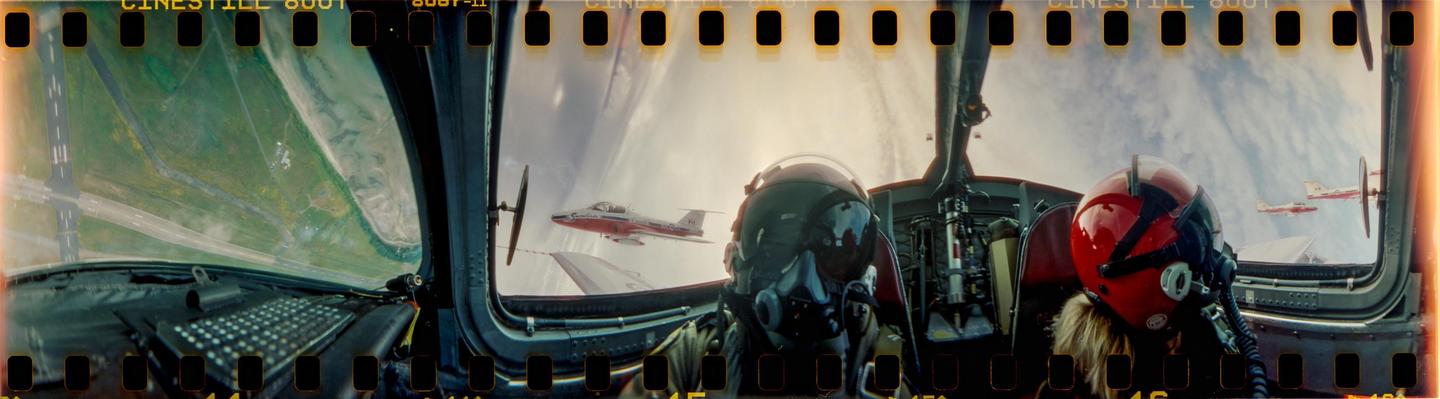 Two pilots in a cockpit fly alongside two other planes over a landscape with an airfield.