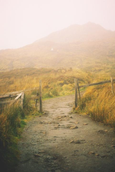 A dirt path leads through a wooden fence towards a hazy mountain.