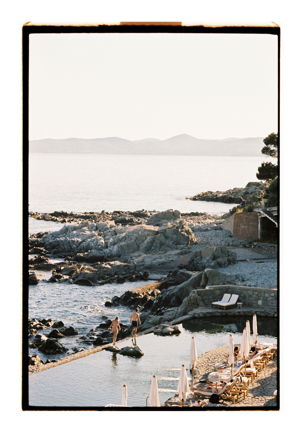 A couple walks along a rocky shore next to a swimming pool with lounge chairs and umbrellas.
