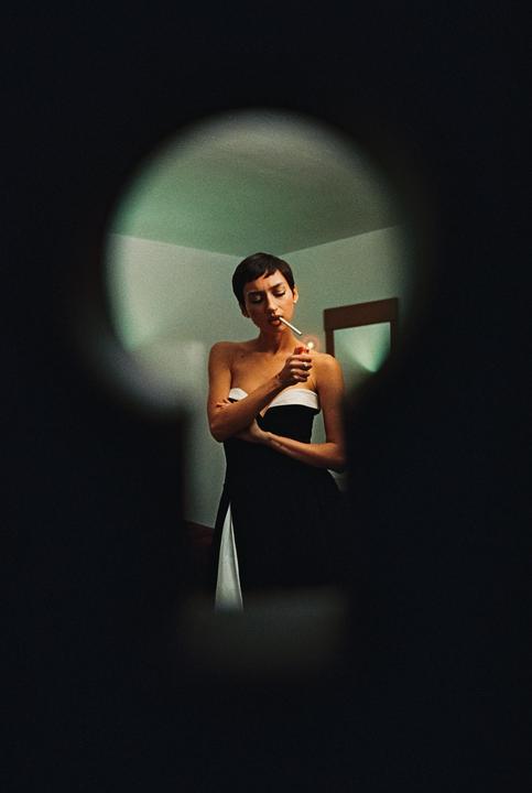 A woman in a strapless dress lights a cigarette, viewed through a keyhole.