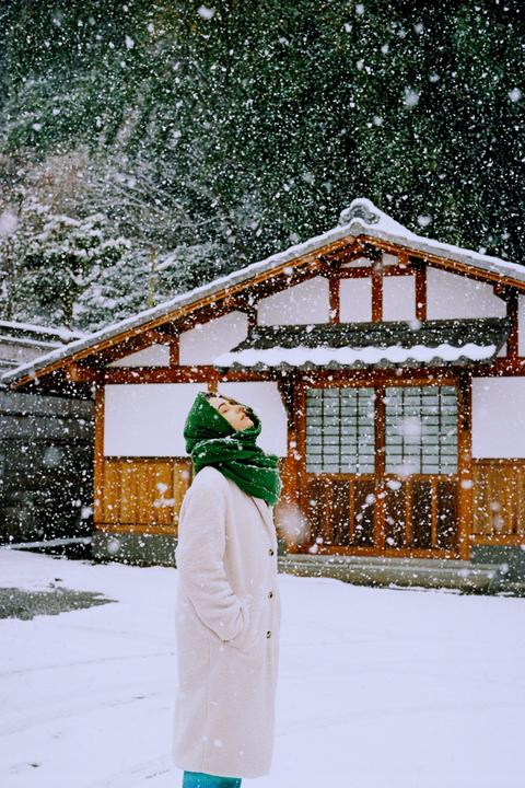 A woman wearing a green scarf and a white coat stands in falling snow, looking up, with a traditional Japanese building in the background.