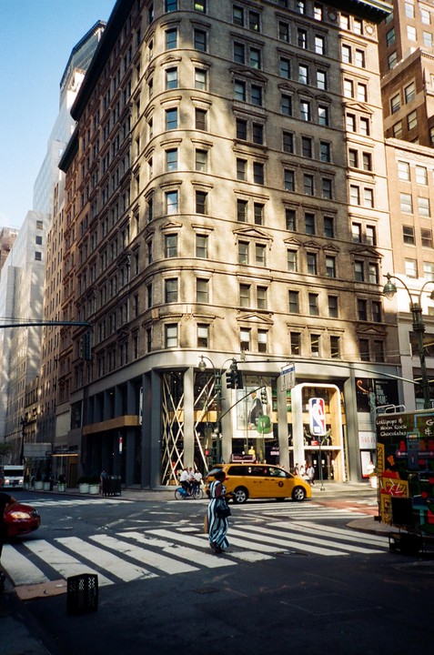 A woman crosses a street in front of a large building with a yellow taxi cab driving by.