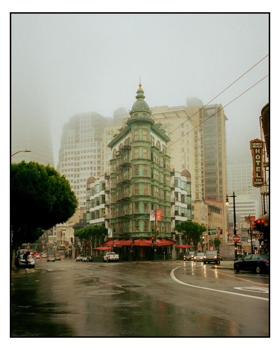 A green building with a domed top stands at a street corner with wet pavement and cars in foggy weather.
