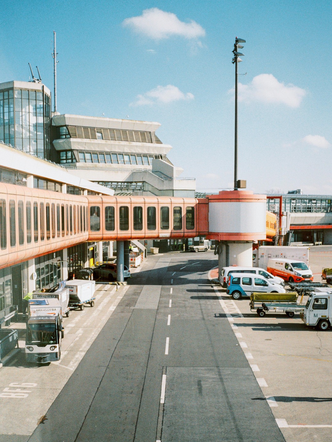 An airport terminal building with a jet bridge extending over an empty tarmac road, with several service vehicles parked nearby under a blue sky with scattered clouds.