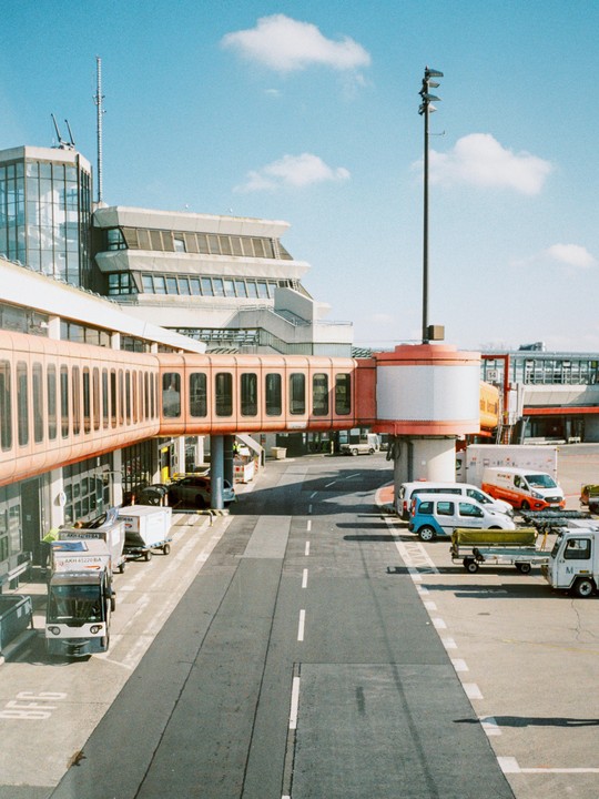 An airport terminal building with a jet bridge extending over an empty tarmac road, with several service vehicles parked nearby under a blue sky with scattered clouds.