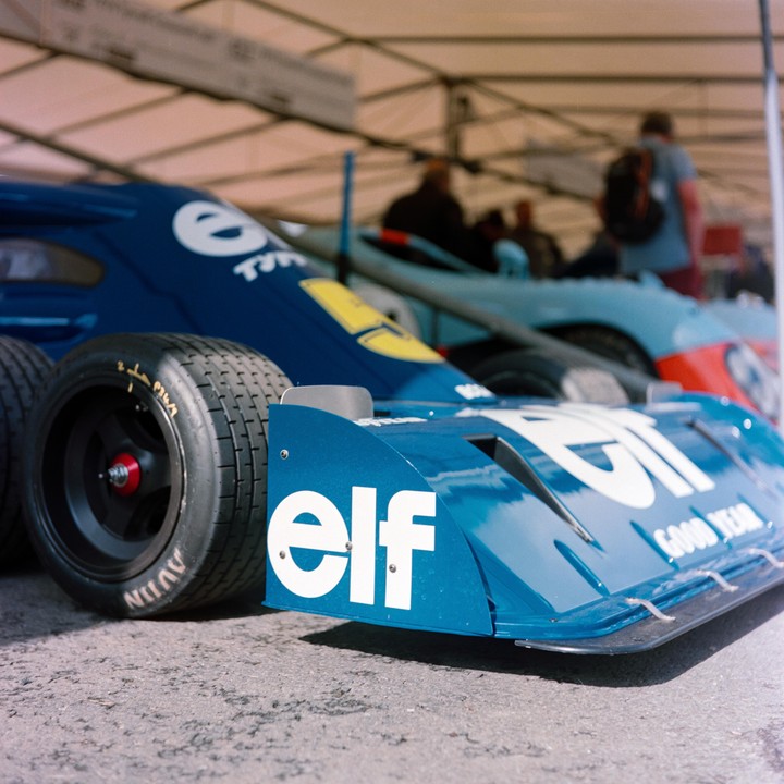 A blue race car with "elf" written on its front wing is parked under a tent, with other cars and people blurred in the background.