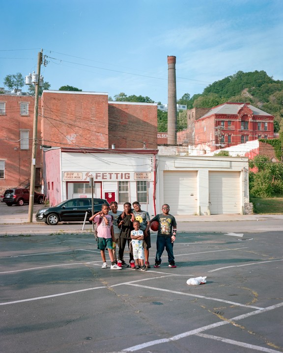 Six boys stand in a parking lot in front of brick buildings and a tall chimney.