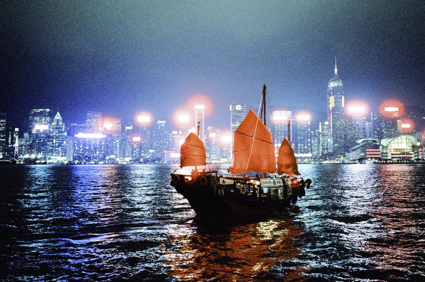A boat with orange sails floats on the water at night with a city skyline in the background.