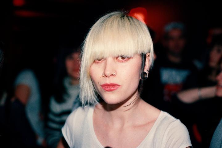 A woman with blonde hair and red lipstick looks directly at the viewer in a dark setting.