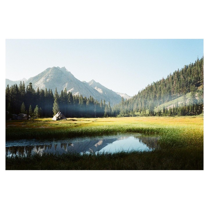 A small pond reflects mountains and a forest in a valley with a grassy meadow.