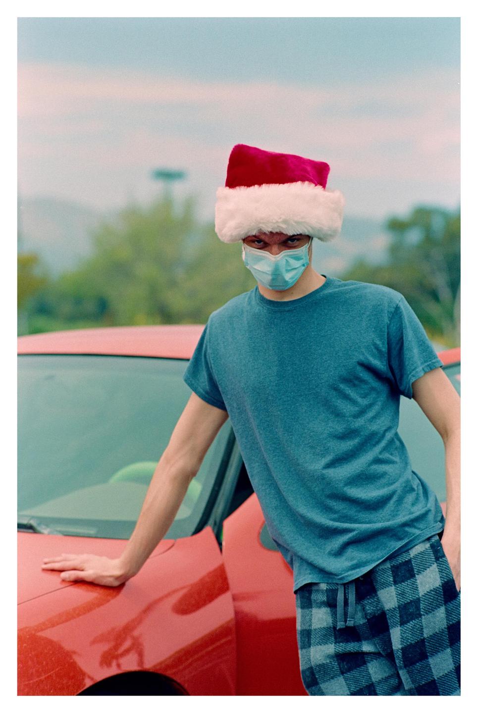 A man wearing a santa hat and a face mask leans against a red car.