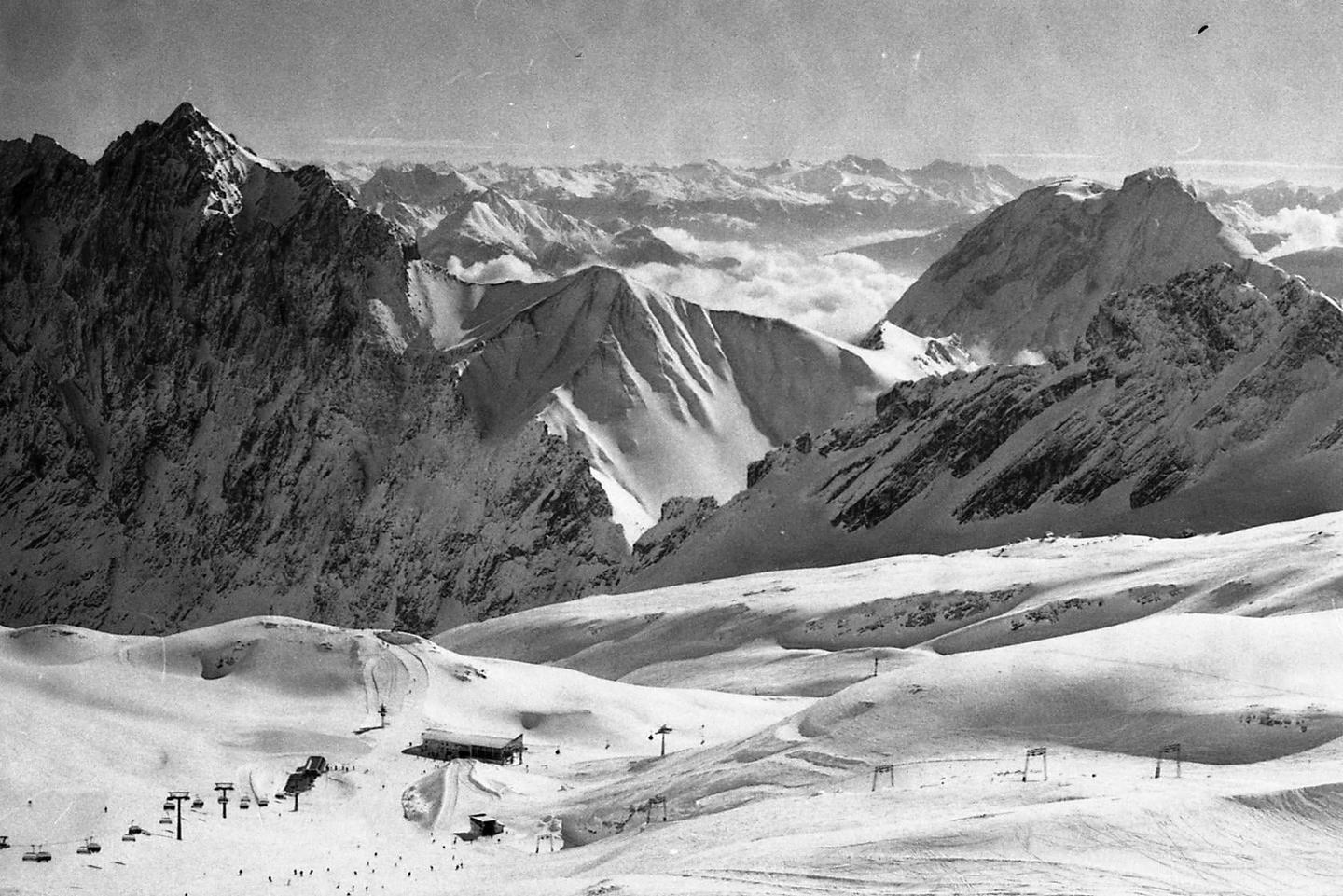 A ski resort with buildings and ski lifts is nestled in a valley surrounded by snow-covered mountains.