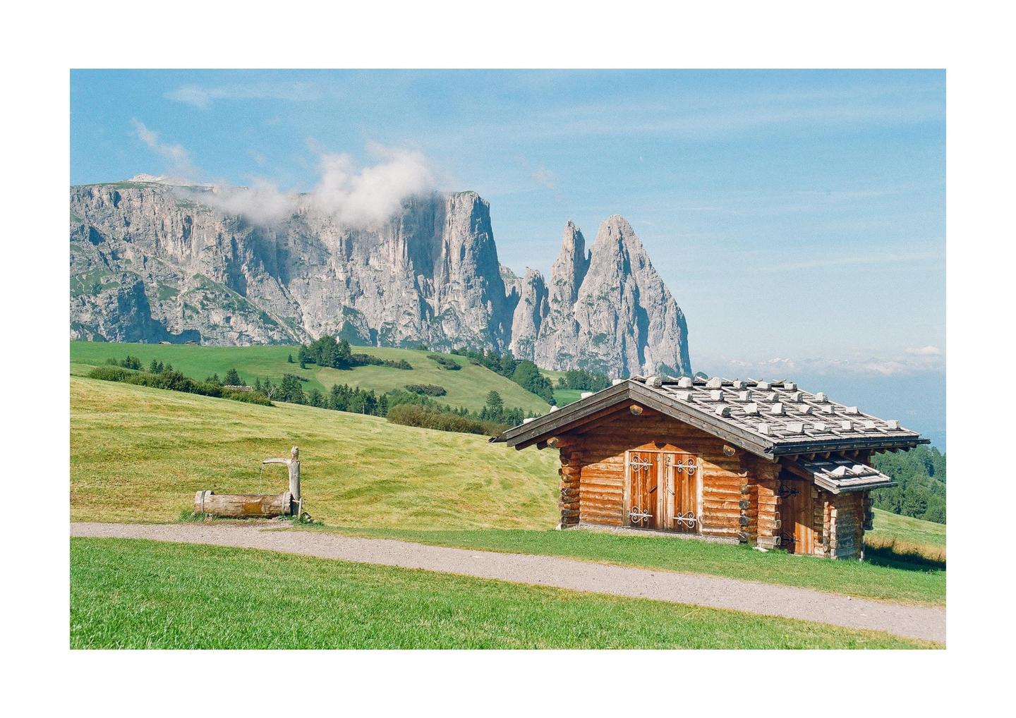 A wooden cabin with a stone roof sits on a grassy hill with a mountain range in the background.
