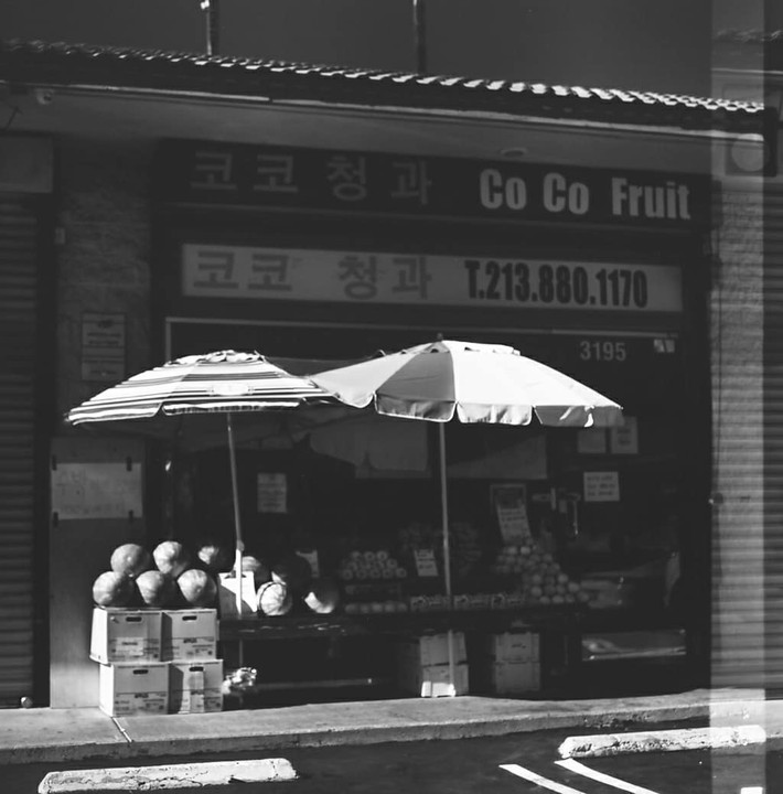 A fruit stand with two umbrellas sits in front of a store with Korean and English text.