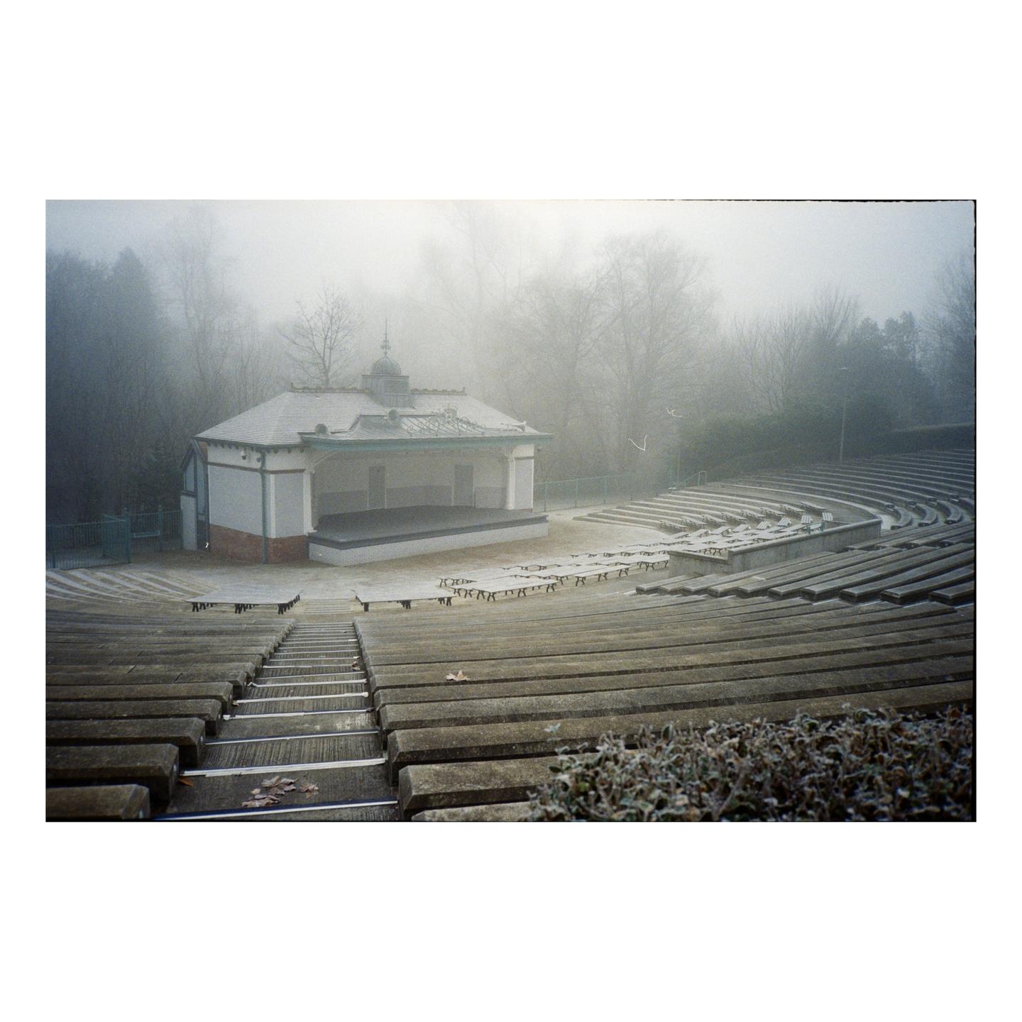 An empty outdoor amphitheater with a bandstand is covered in a light dusting of snow on a foggy day.