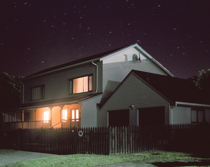 A house with illuminated windows stands under a night sky filled with star trails.