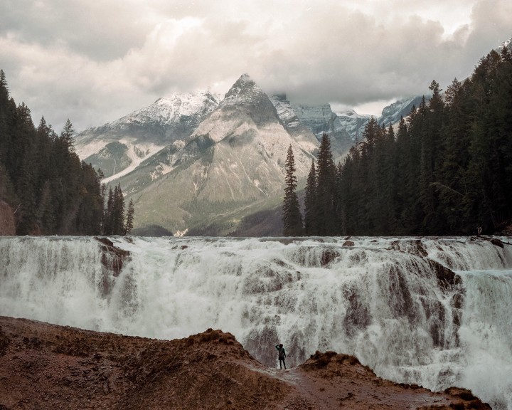 A person stands on a rocky outcrop overlooking a wide waterfall with a forest and snow-capped mountains in the background under a cloudy sky.