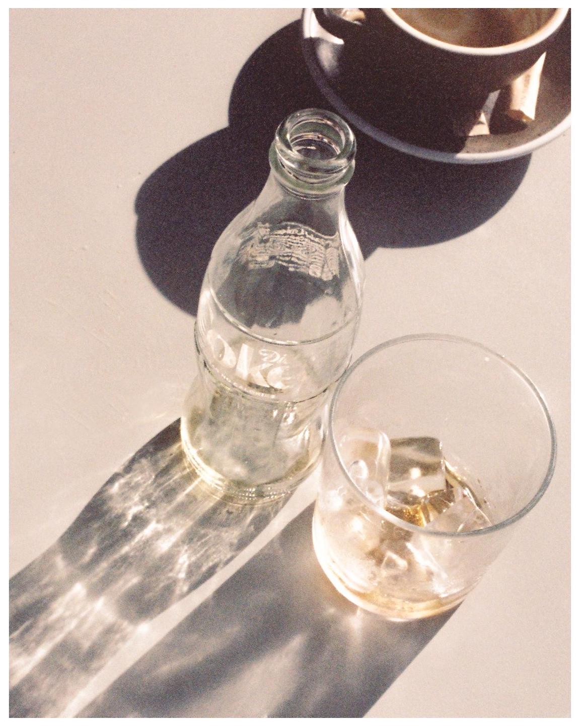 A glass bottle, a glass with ice, and a cup are on a table with strong shadows.