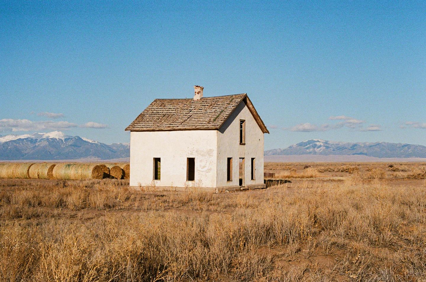 An abandoned house stands in a dry field with mountains in the distance.