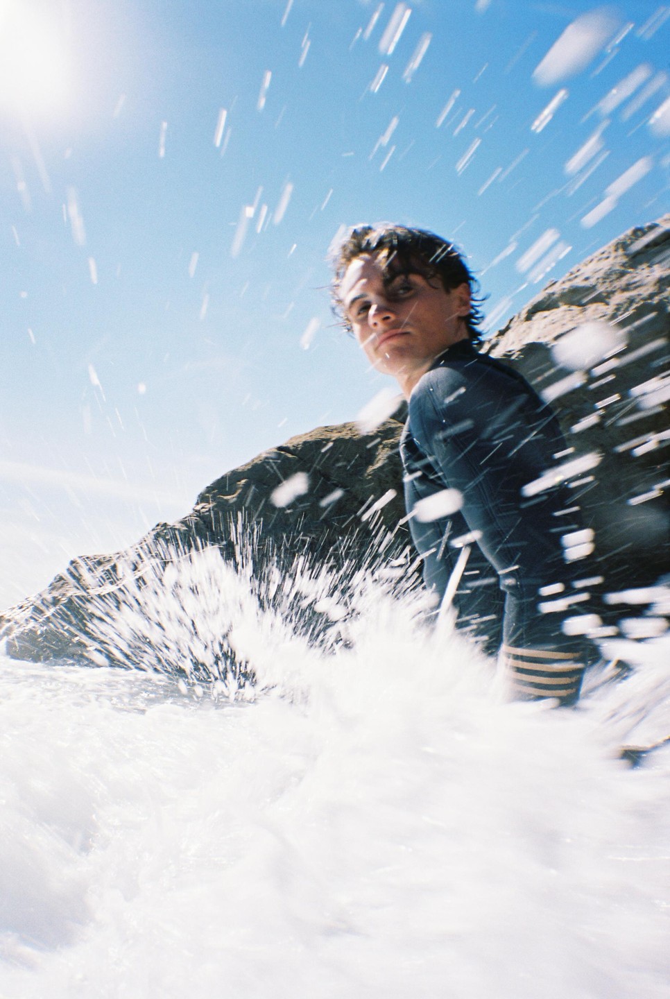 A man in a wetsuit stands in the ocean with water splashing around him and a rocky cliff in the background.