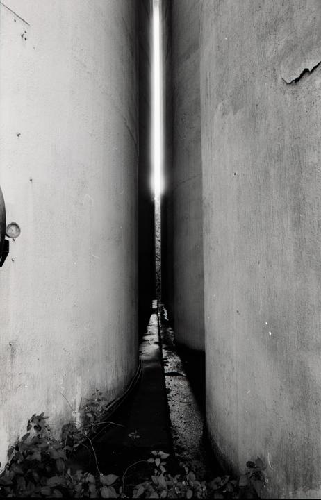 A narrow alleyway between two tall buildings leads to a bright light at the end.