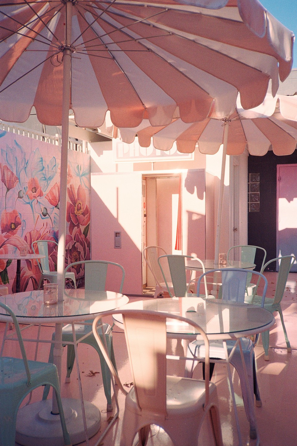 A patio with tables and chairs is shaded by two large umbrellas, with a pink building and a floral mural in the background.