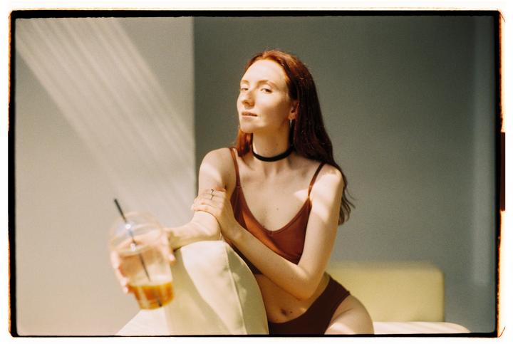 A woman in a brown top and underwear sits on a couch, holding a drink with ice and a straw.