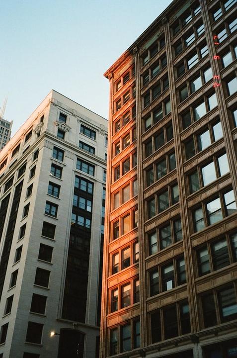 Two tall buildings stand against a clear sky, with one building illuminated by golden light.