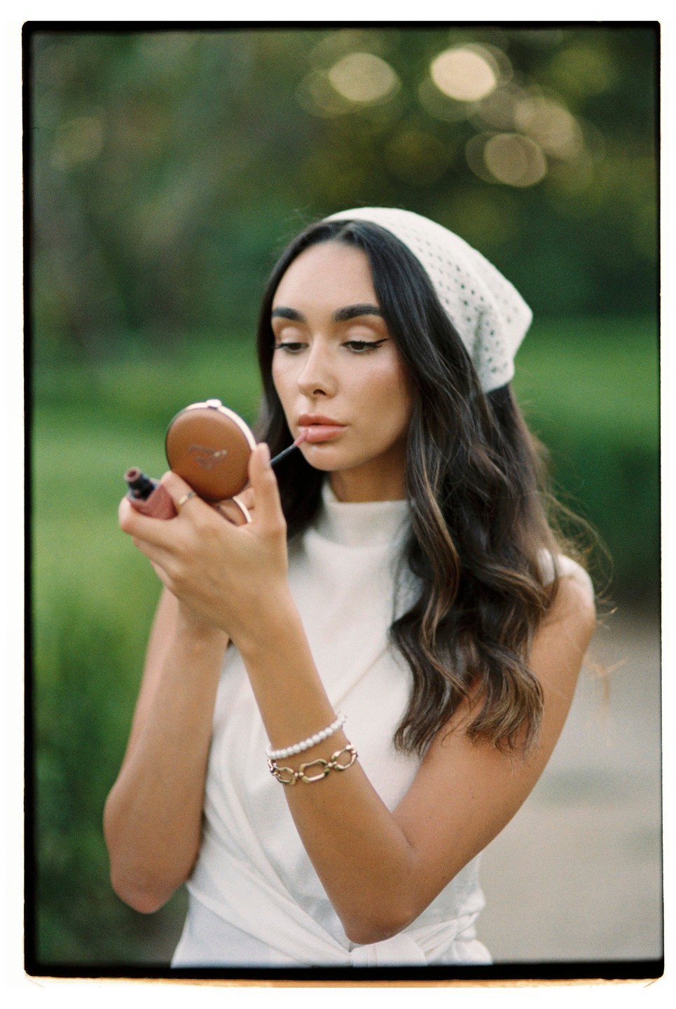 A woman with long dark hair and a white headscarf applies lipstick while looking into a compact mirror.