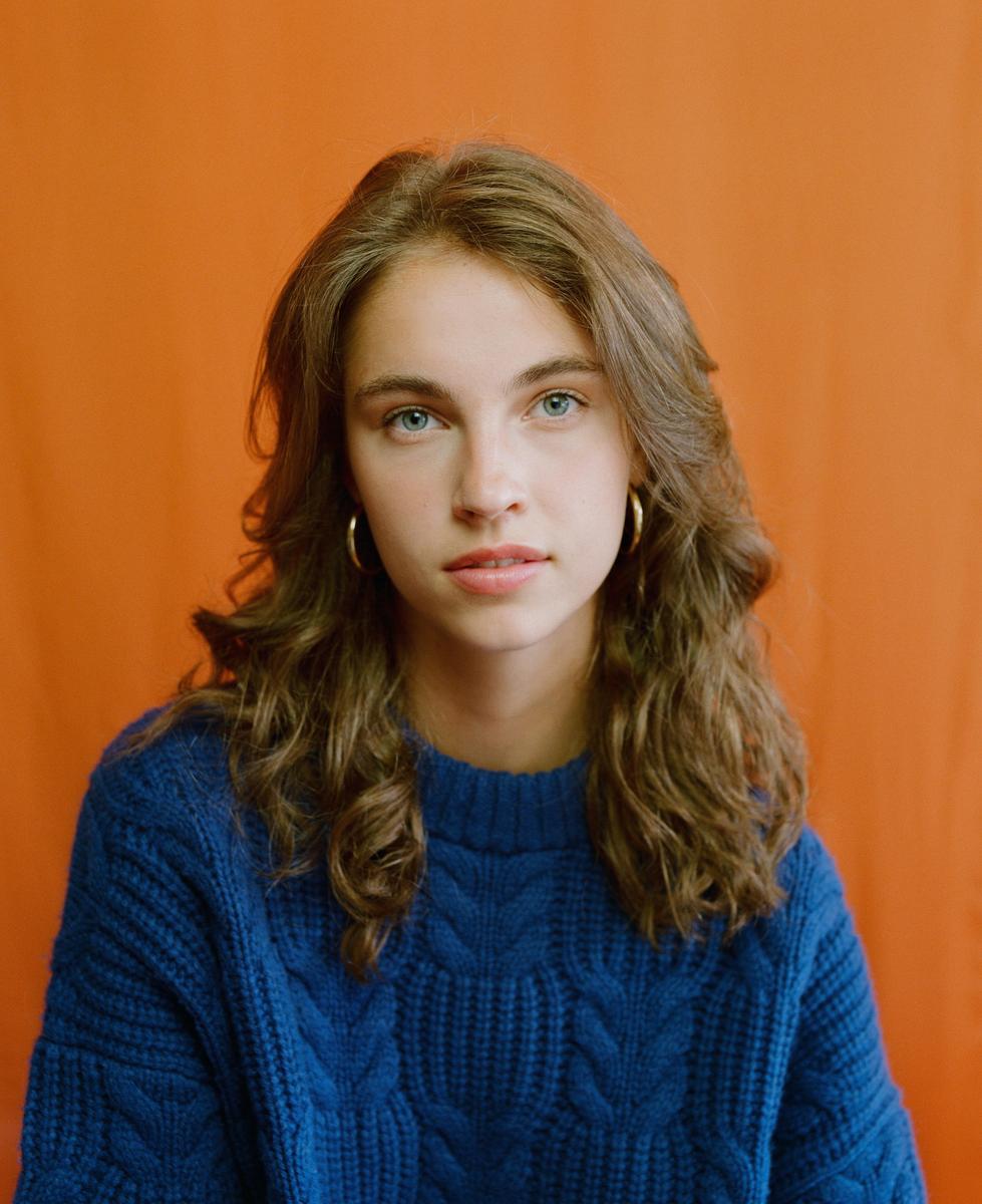 A young woman with long, wavy hair and a blue cable-knit sweater looks directly at the viewer against an orange background.
