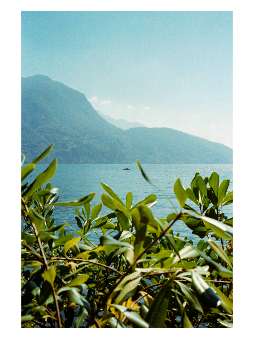 A small boat is visible on a lake with mountains in the background, framed by green foliage in the foreground.
