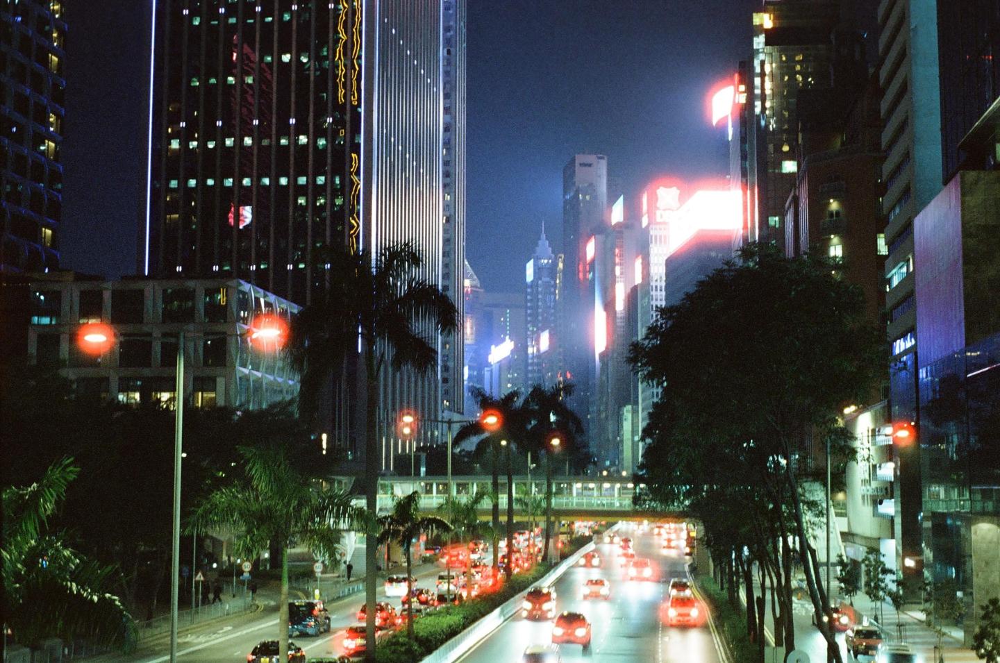 A busy city street at night is filled with cars and surrounded by tall buildings and palm trees.