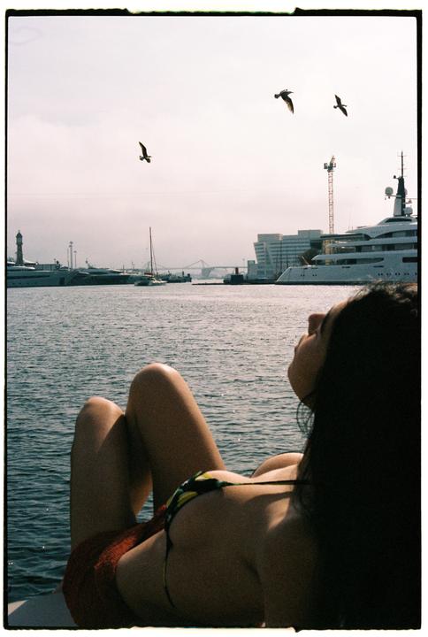 A woman in a bikini lies on a boat looking at the water with birds flying overhead and a city in the background.