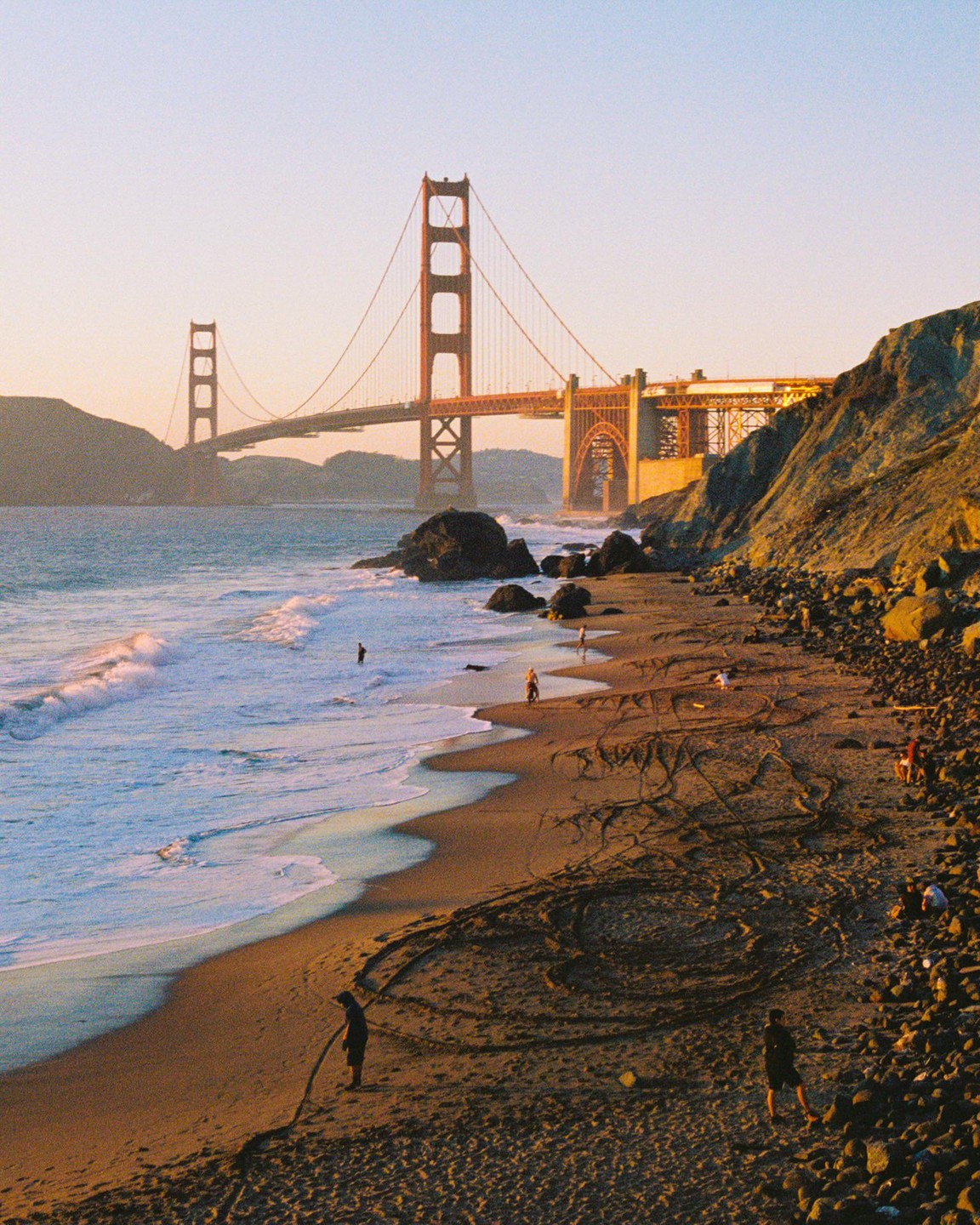 The Golden Gate Bridge is visible in the distance from a beach with people and waves.