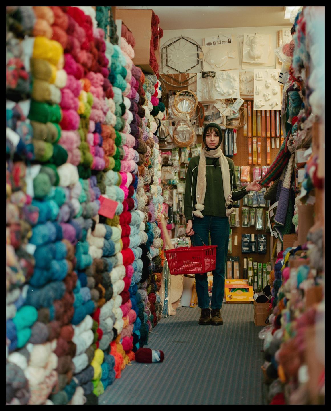 A woman stands in a narrow aisle of a yarn shop, holding a red basket.