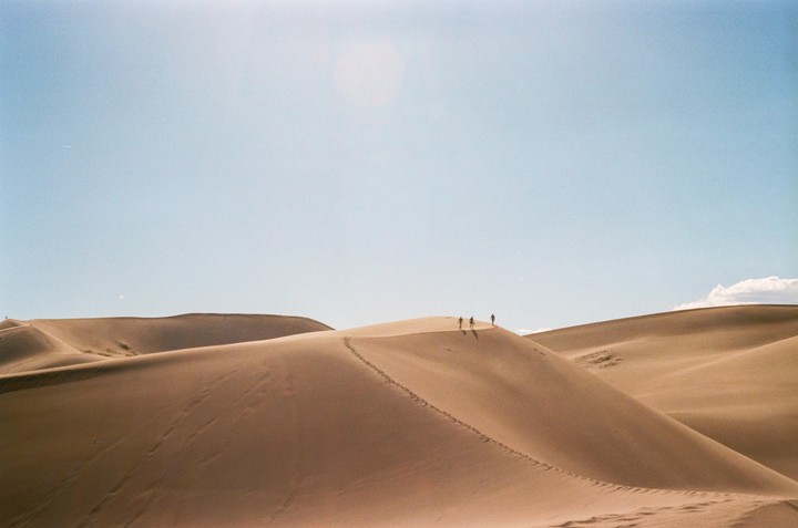 Three people walk along a ridge of sand dunes under a clear sky.