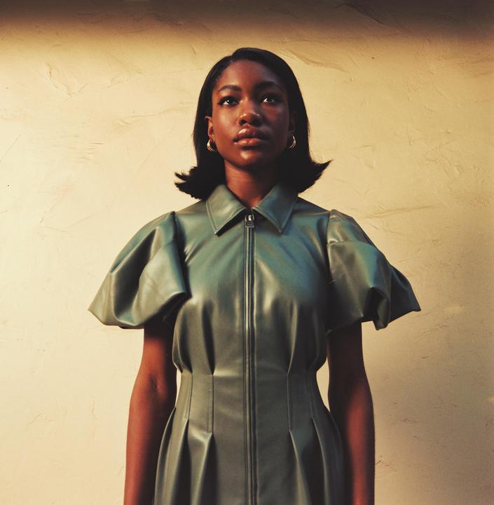 A woman in a green dress with puffed sleeves stands against a textured wall.