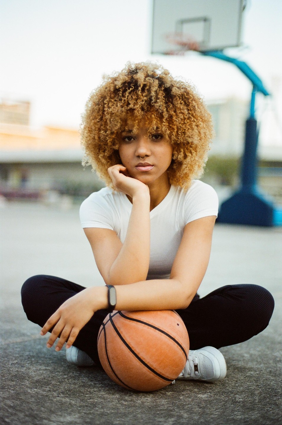 A young woman with curly hair sits on a basketball court with a basketball in front of her.